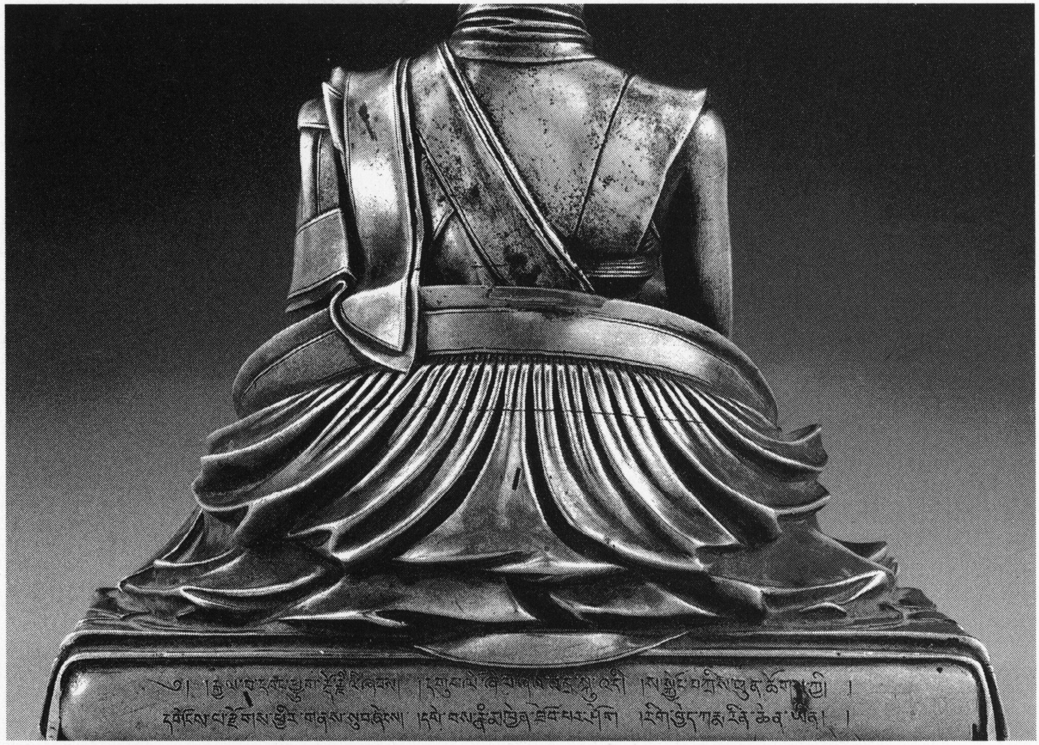 Back view of statuette depicting lama wearing pleated robe; two lines of Tibetan text inscribed at base