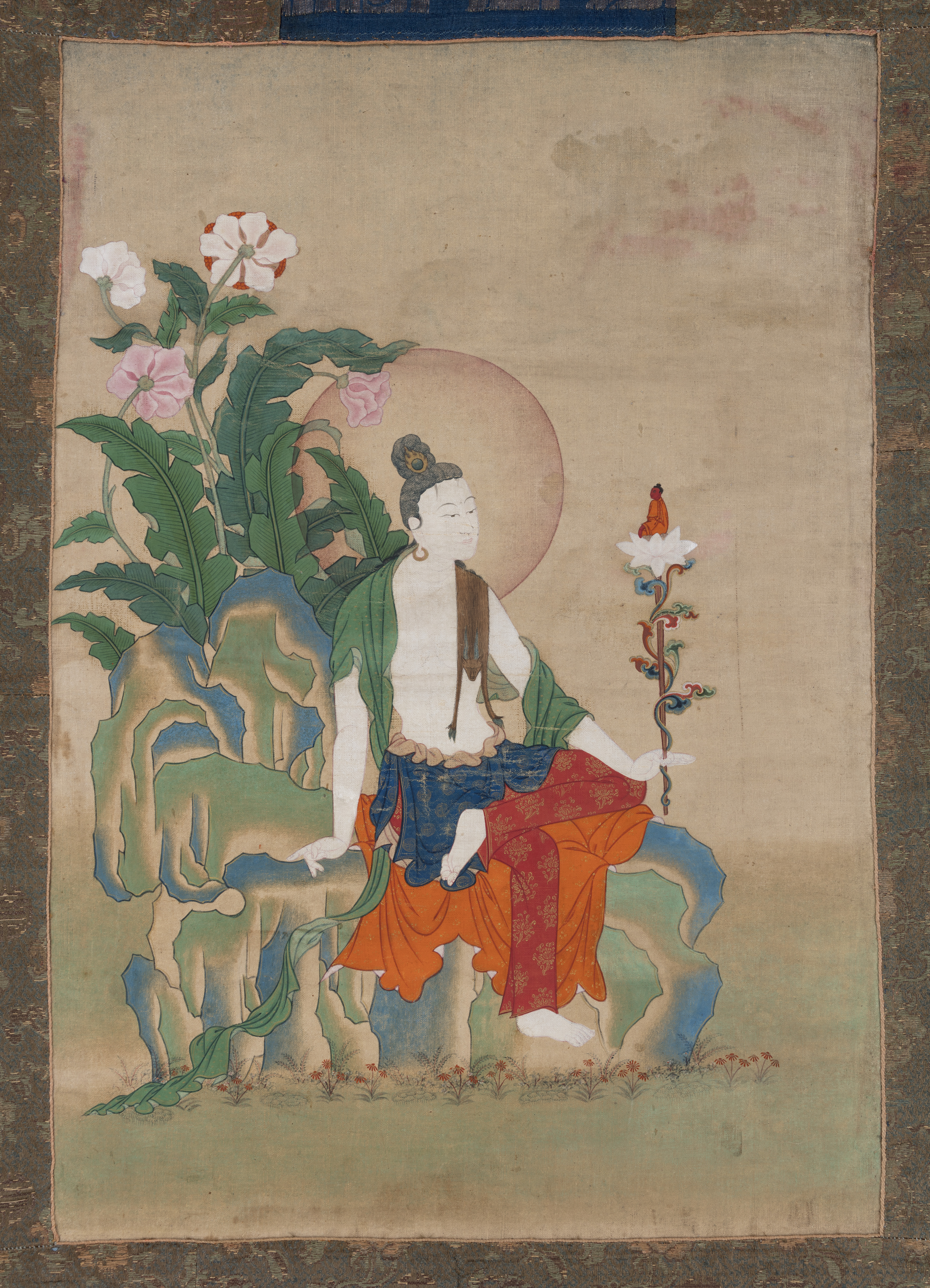 Bodhisattva dressed in sumptuous dhoti holds blossom in right hand while seated on rocky outcropping