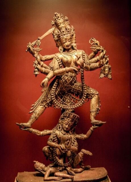 Statue depicting fearsome many-armed goddess standing in dynamic pose atop two figures (one kneeling, one supine)