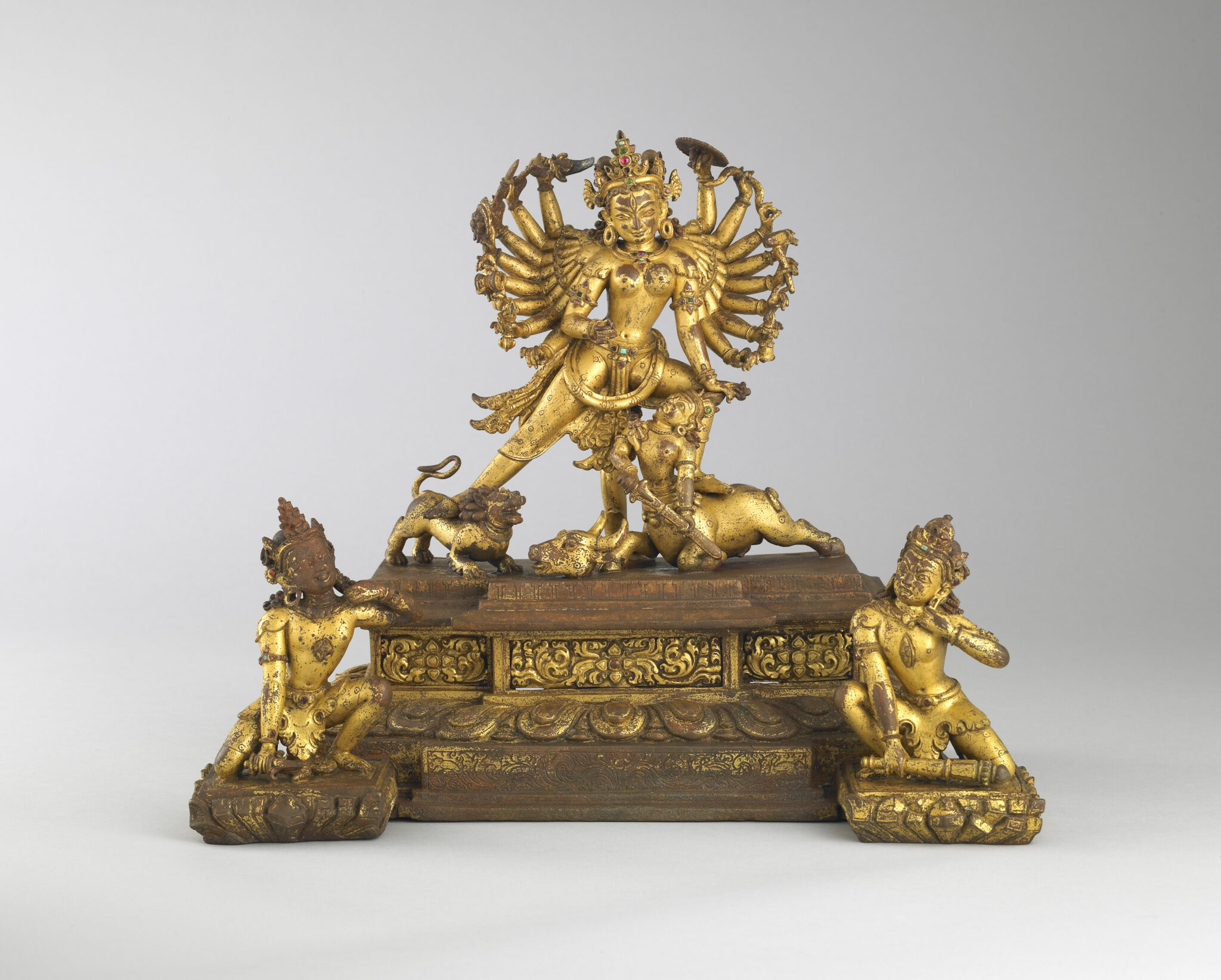 Sculpture depicting golden, many-armed goddess and animals atop wooden plinth flanked by kneeling attendants
