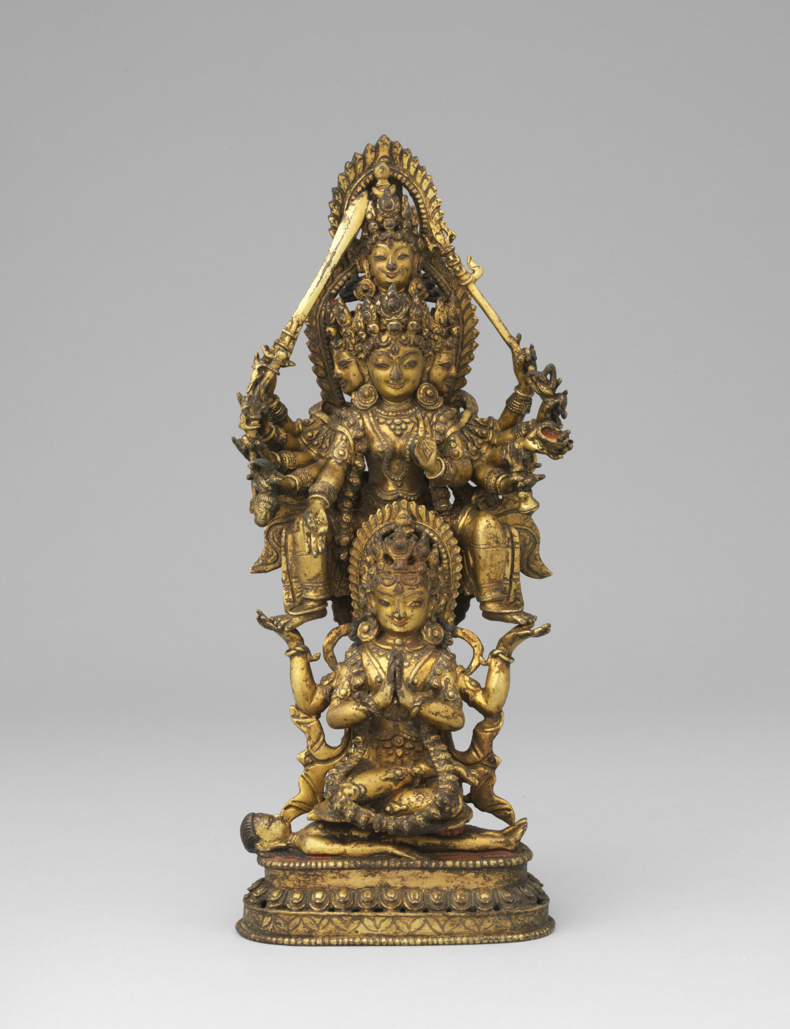 Golden statuette depicting goddess composed of two figures: below, seated figure; above, many-headed and -armed figure