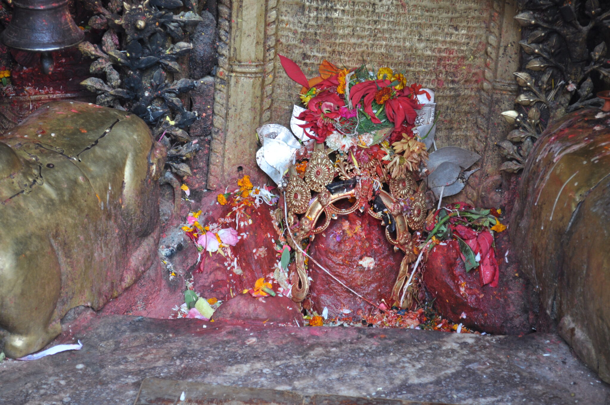 Three sacred boulders painted red and strewn with flowers; middle boulder adorned with gold and silver crown