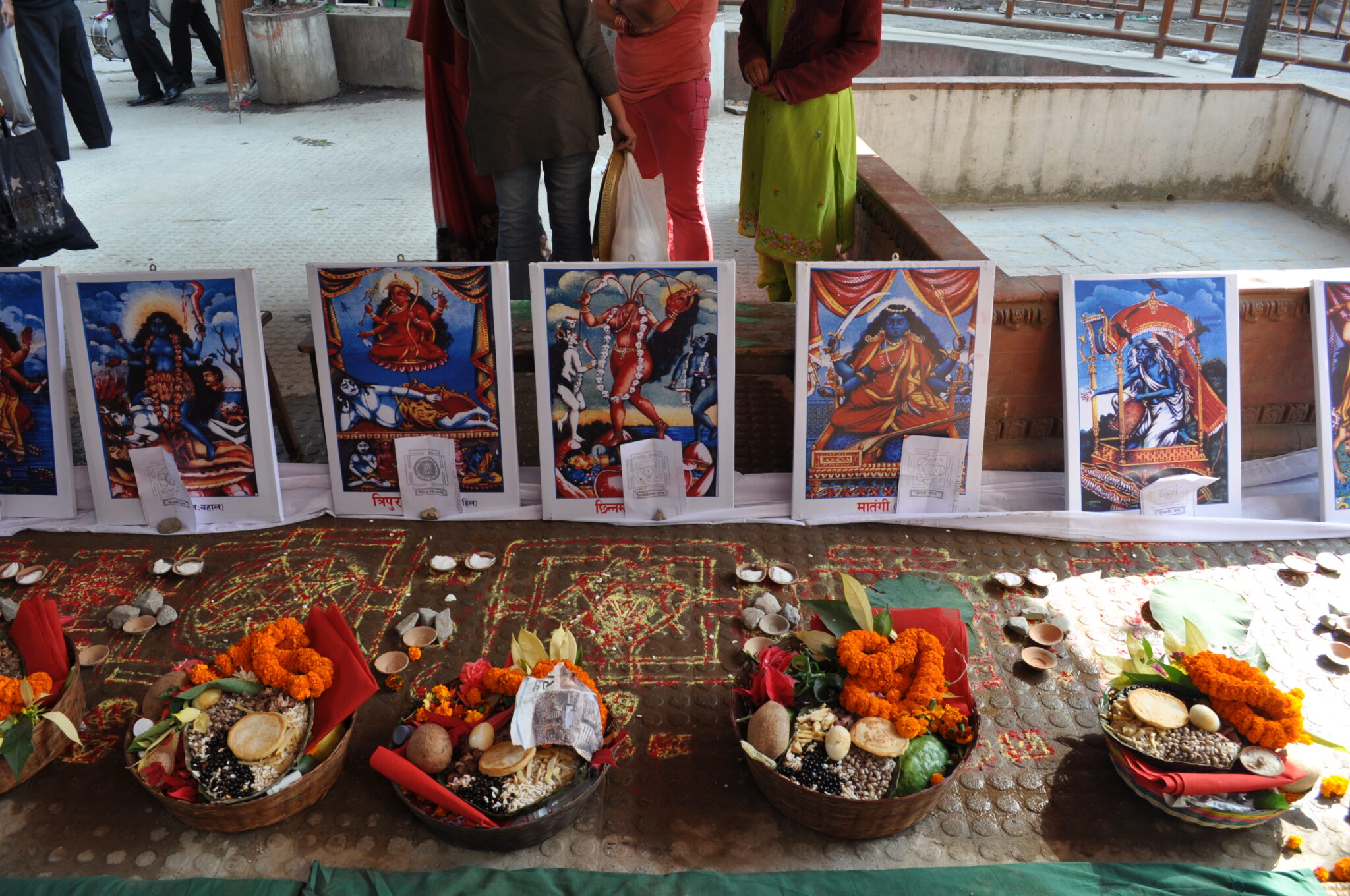 Arrangement of postcard-sized prints depicting deities before bowls brimming with offerings atop embroidered textile