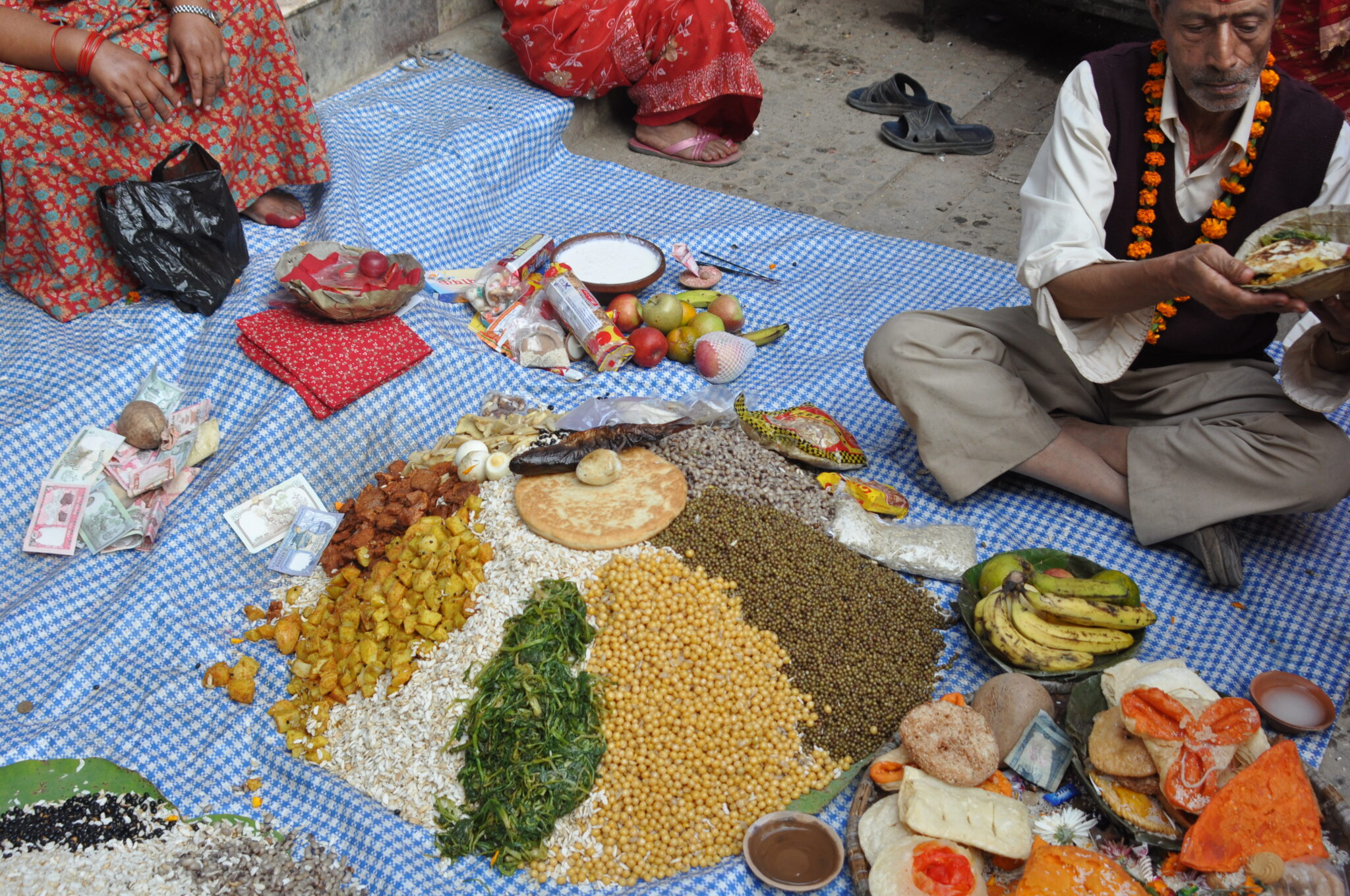 Arrangement of grains, vegetables, and meats surrounded by worshippers and other offerings atop blue gingham cloth