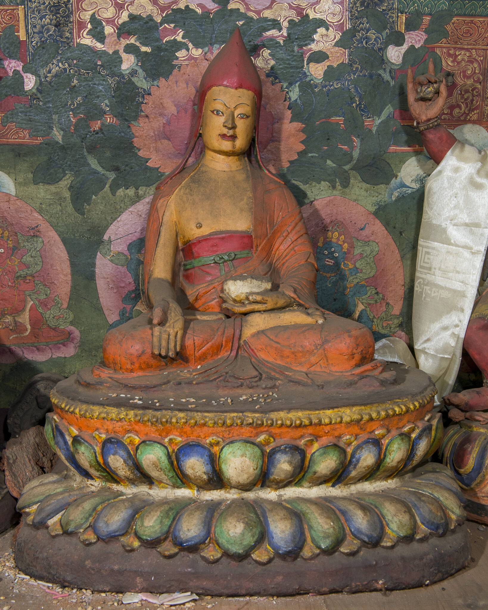 Sculpture depicting figure wearing saffron robe and pointed red headdress seated on lotus pedestal before mural