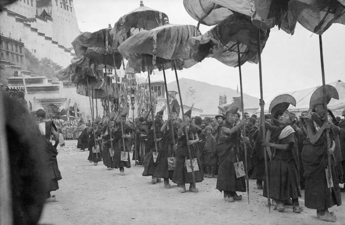Black and white photograph of monks carrying parasols, billowing in wind, at base of hilltop palace