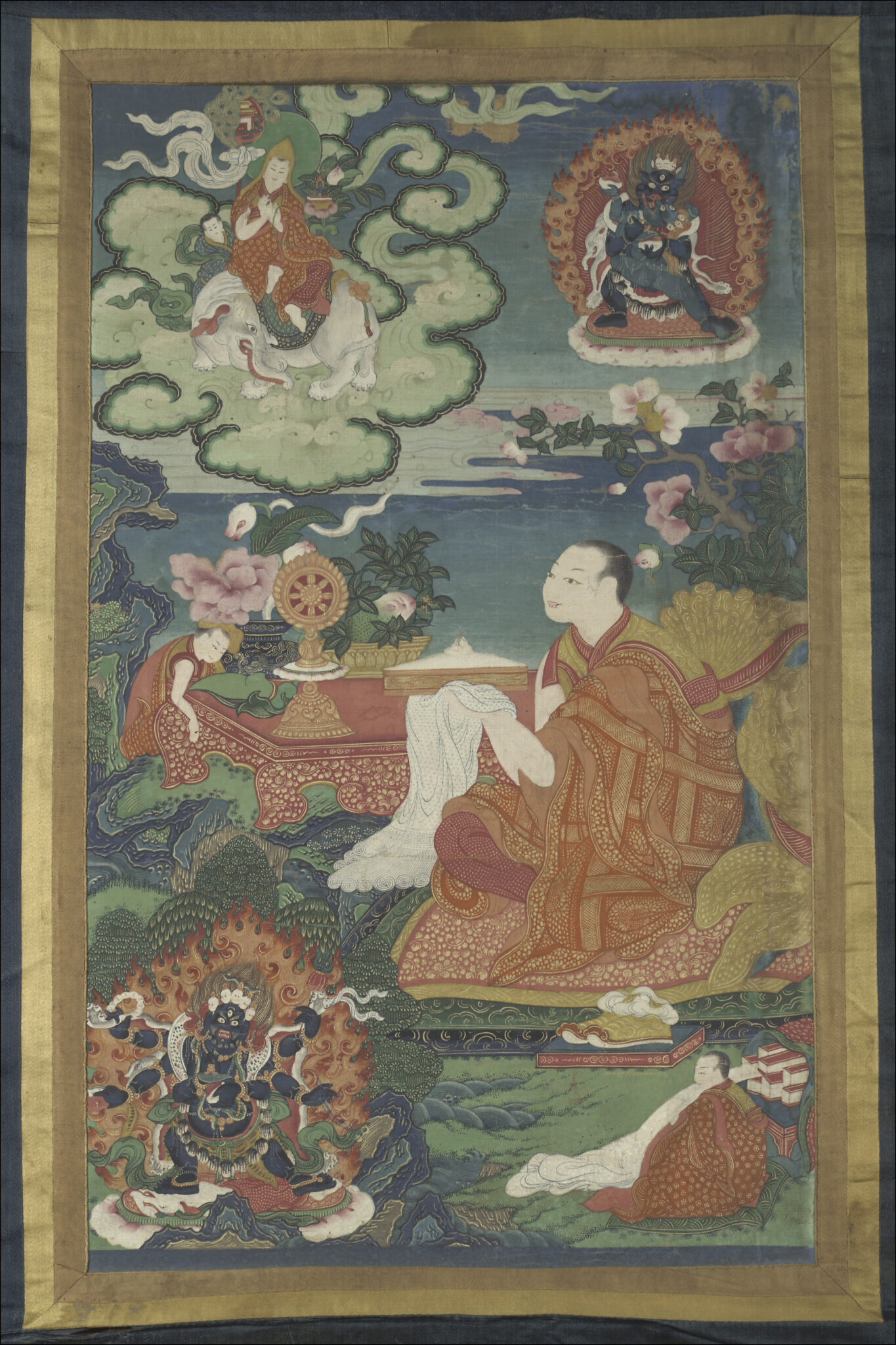 Lama wearing saffron patchwork robe, holding white prayer scarf, gazes at table adorned with implements and blossoms