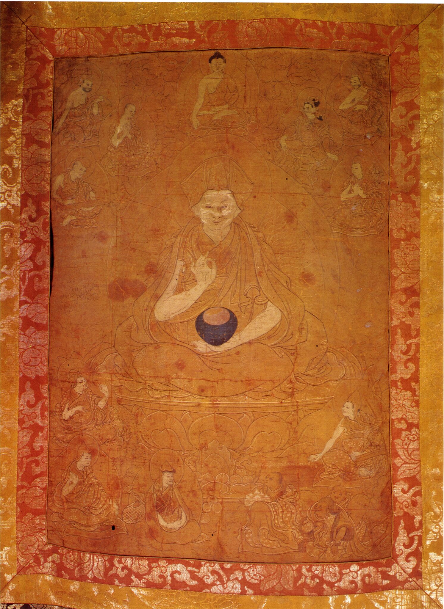 Saffron-colored painting mounted on orange and yellow brocade depicting seated Lama surrounded by small figures
