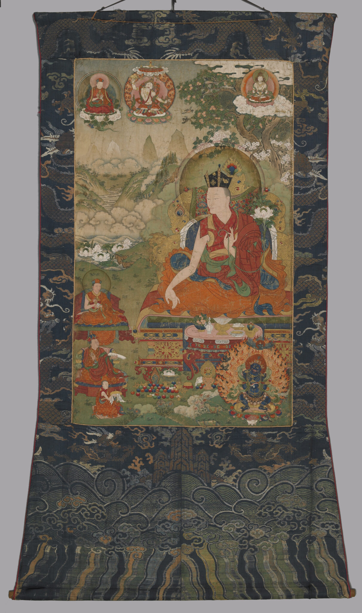 Painting, mounted on blue brocade featuring dragon motif, depicting lama seated before attendants in mountain landscape