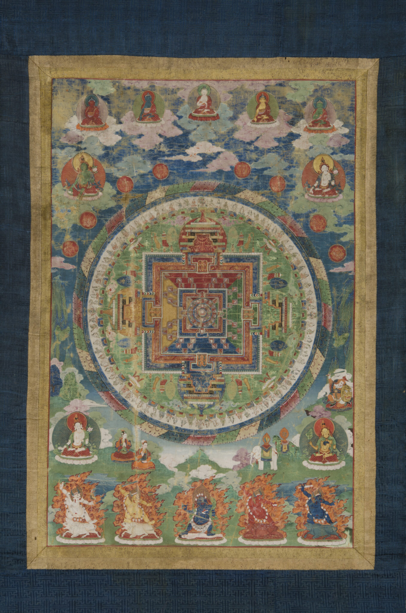 Mandala mounted on blue and gold textile border featuring deity portraits at bottom and background landscape