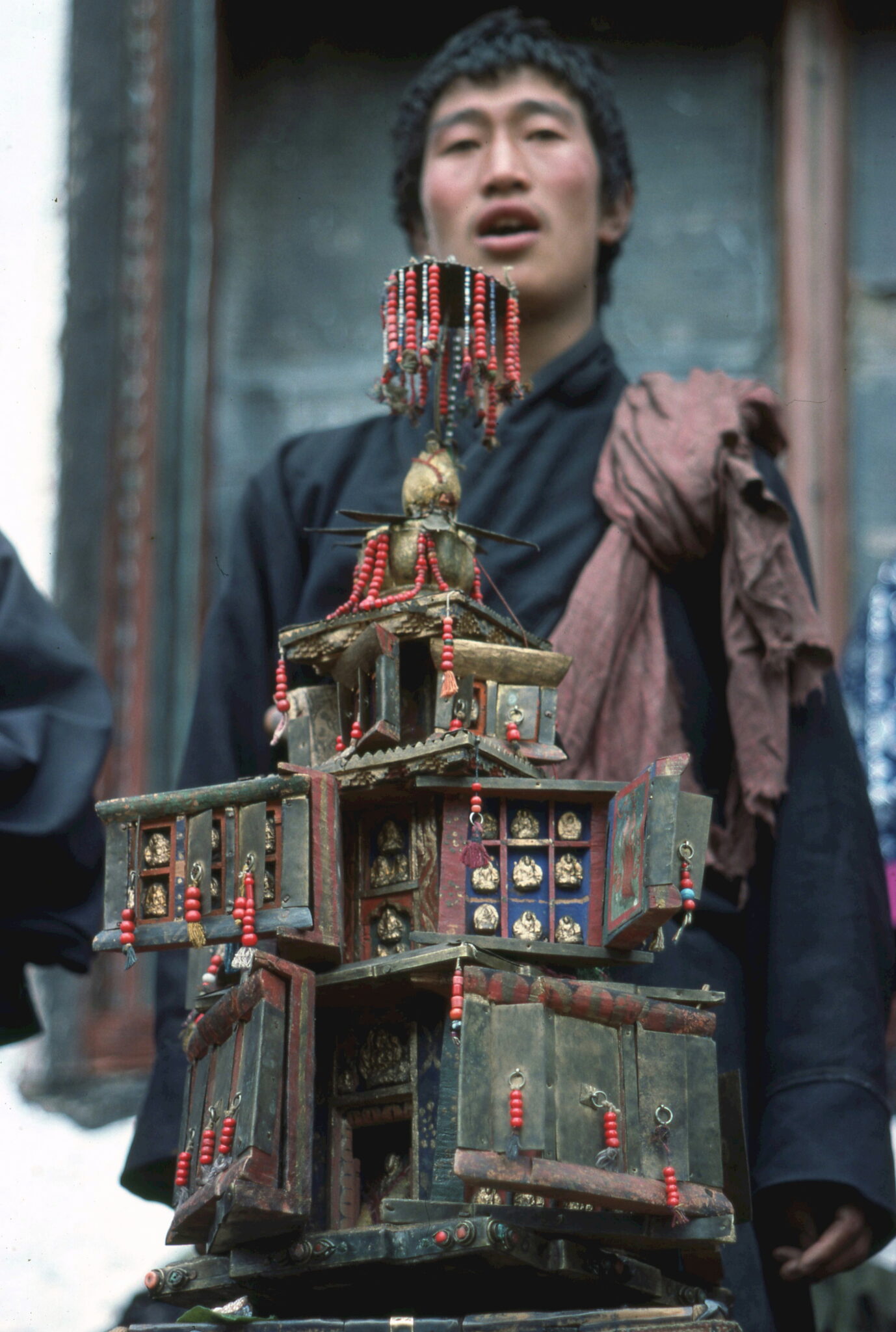 Man stands behind religious implement in the form of tiered building decorated with red beads