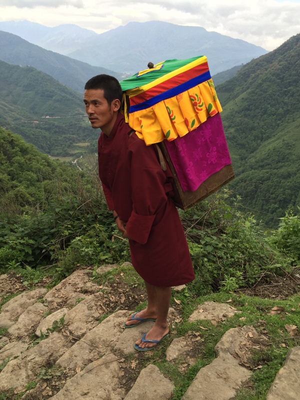 Man dressed in red tunic carries box with colorful textile cover on his back through mountain valley