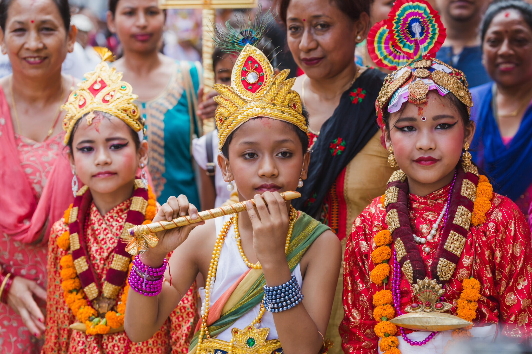 Child wearing golden headdress plays flute while flanked by two children in colorful garments and headdresses