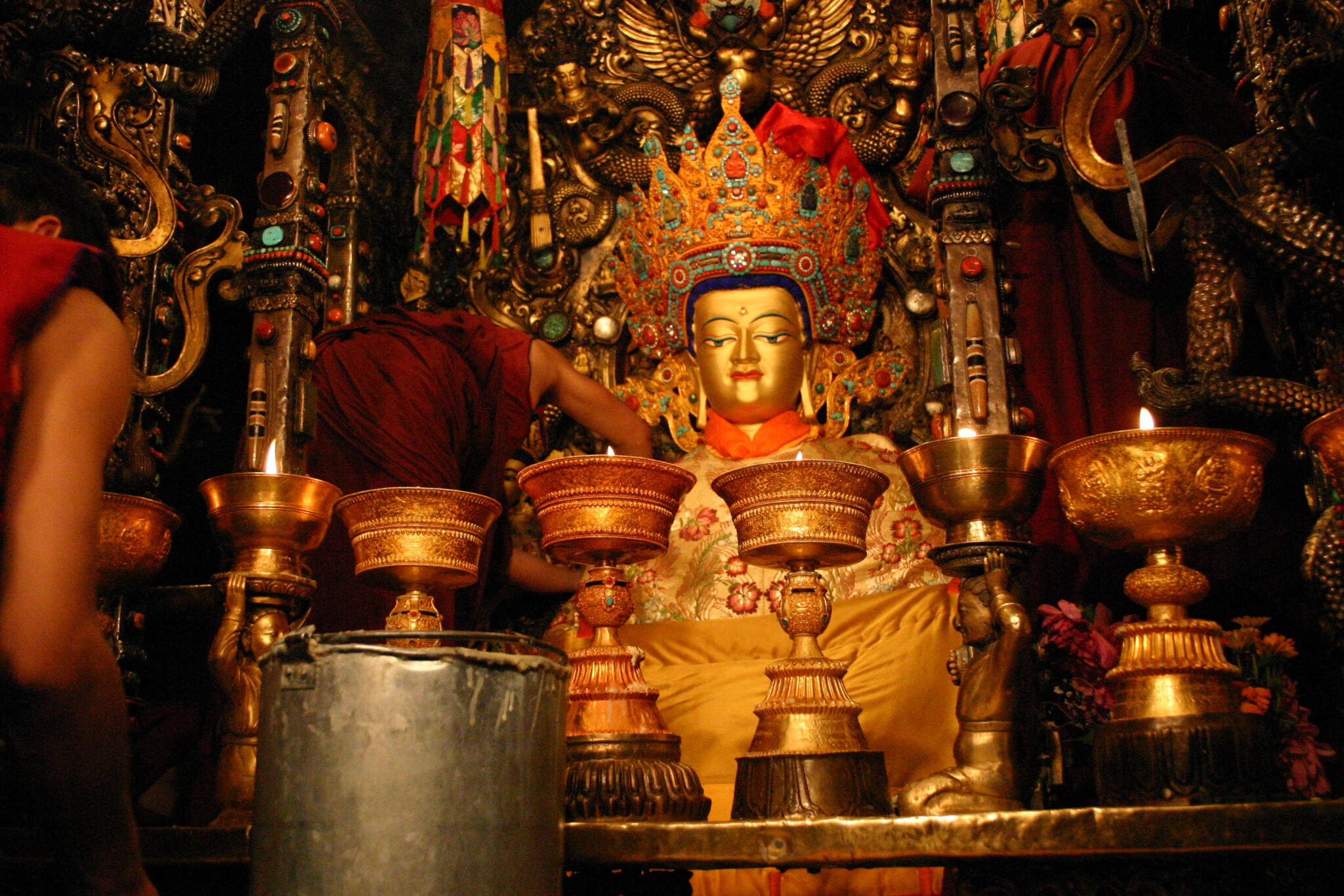 Monks approach golden statue of Buddha from the left; scene is viewed through row of butter lamps
