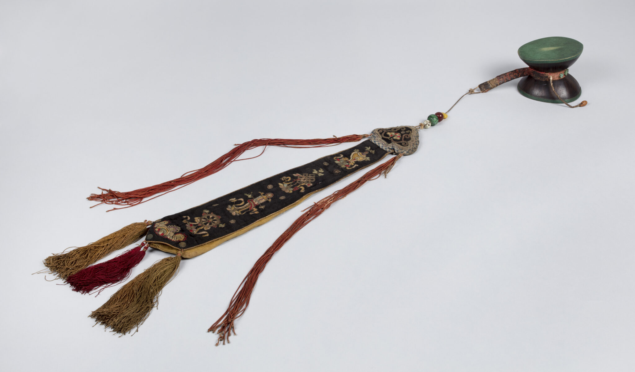 Small, green, double-headed drum attached with cord to long tassel embroidered with religious motifs
