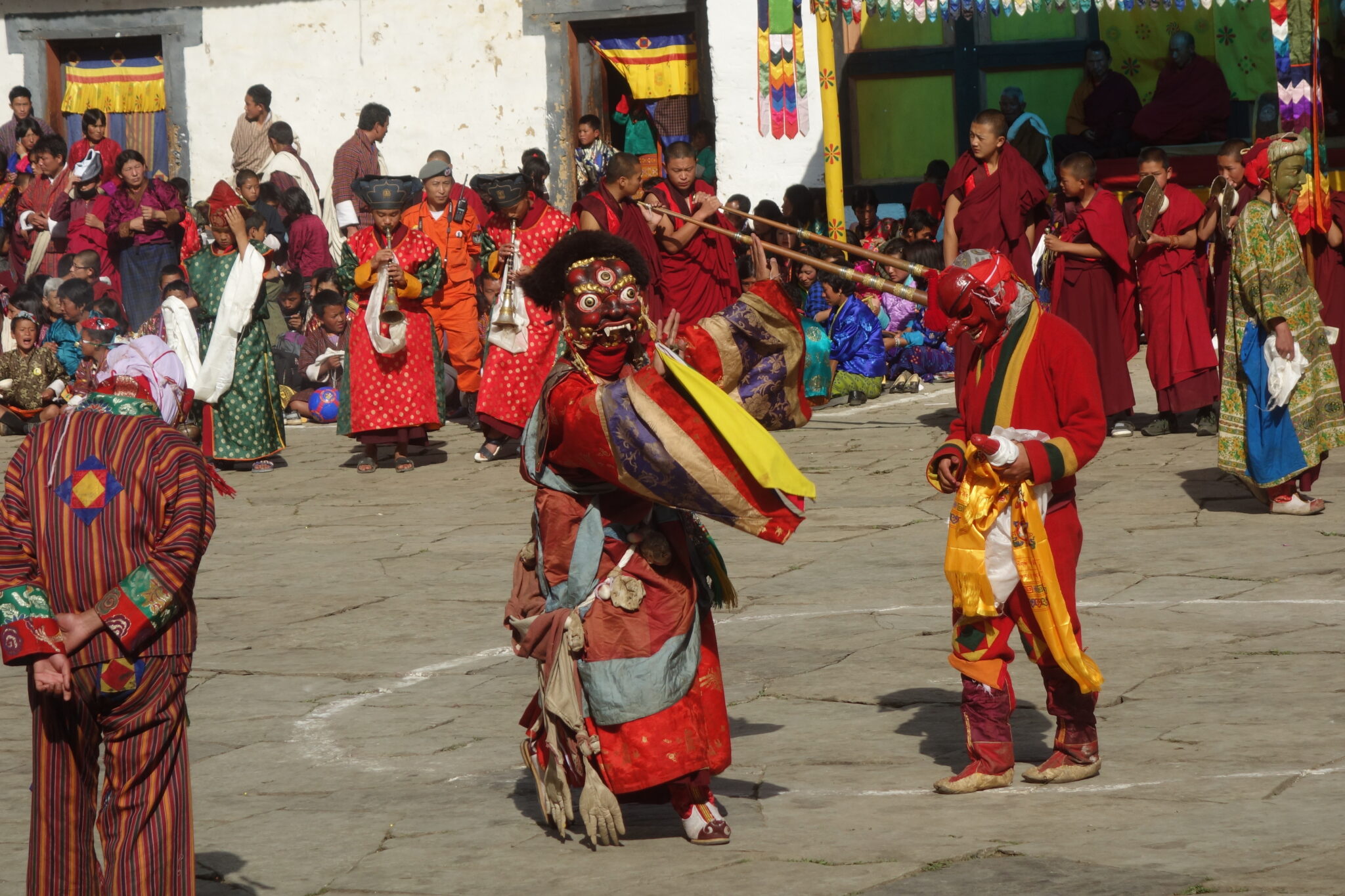 Two performers dressed in red masks and flowing garments dance before musicians in forecourt of building