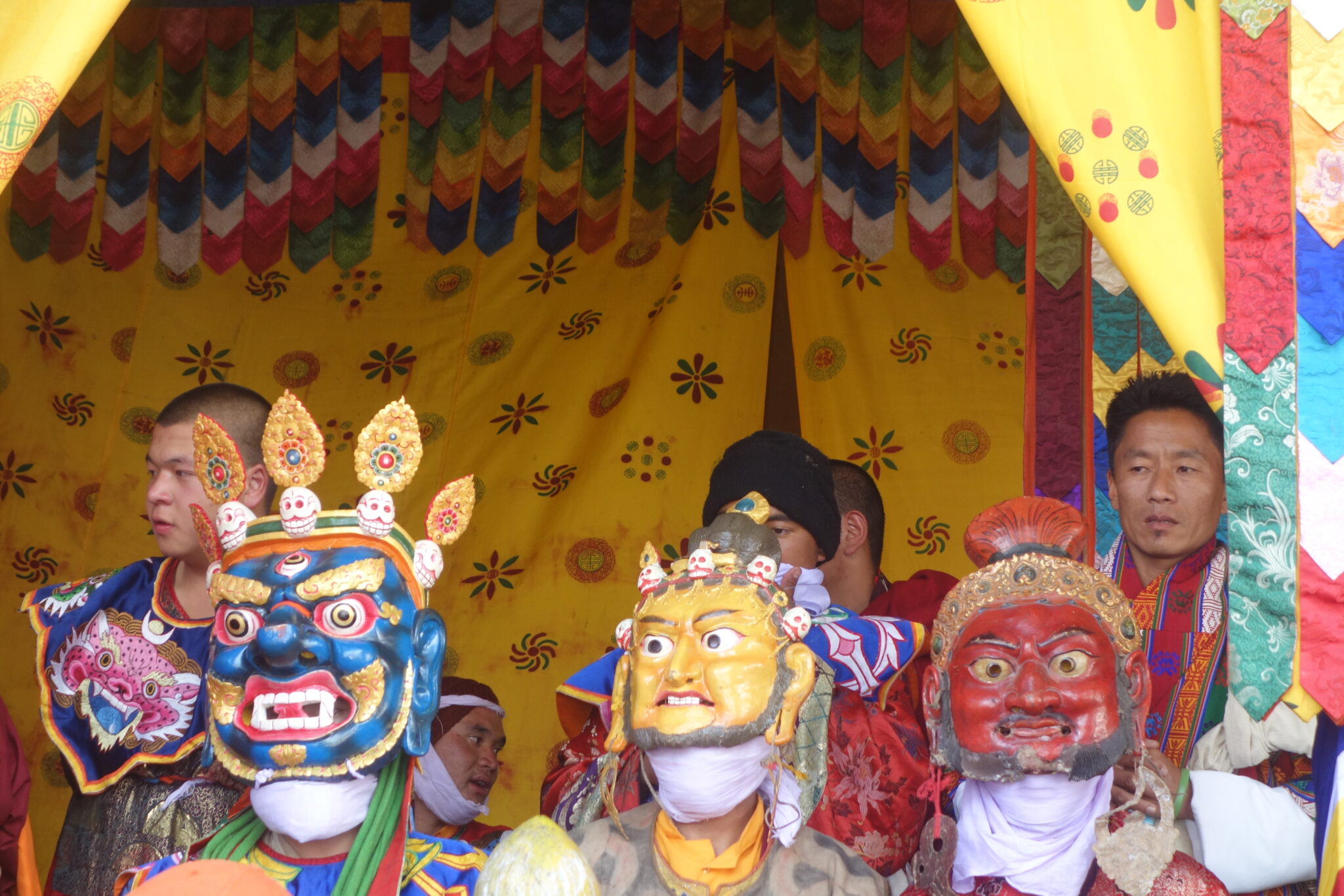 Three performers wearing masks in blue, yellow, and red pictured before structure festooned with saffron textile and banners