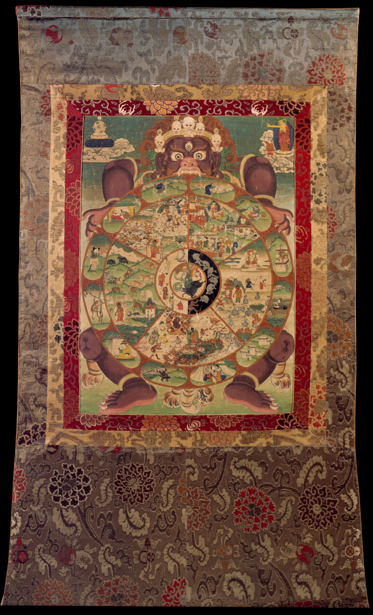 Painting mounted on brocade depicting wheel divided into multiple scenes held by wrathful deity