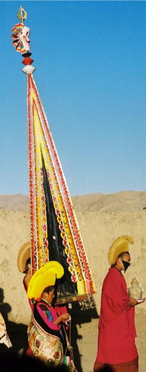 Lamas carry tall, triangular sculpture in black, yellow, and red with stylized skull affixed to point