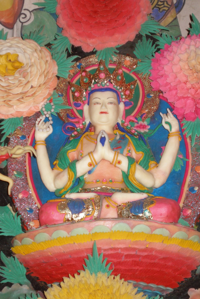 Colorful butter sculpture depicting four-armed, crowned Bodhisattva seated underneath trio of chrysanthemums
