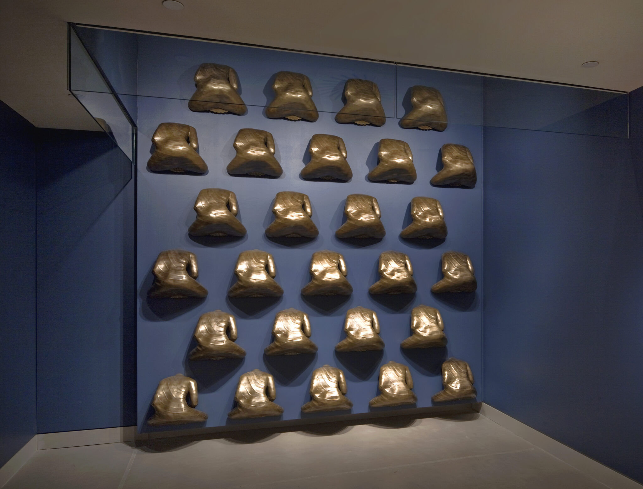 27 golden Buddha statues, turned away from viewer, mounted in six rows with backsides protruding from blue wall