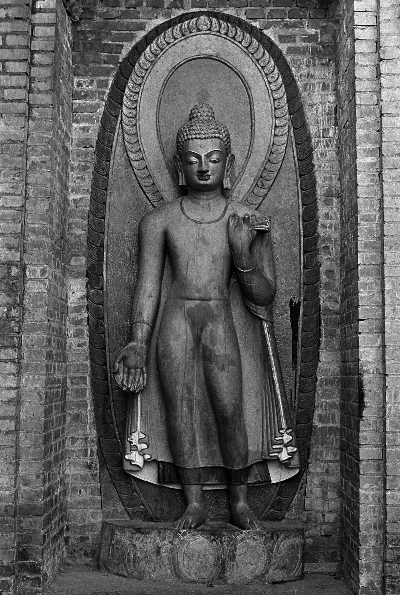 Black and white photograph of Buddha standing with right hand raised in mudra situated in brickwork niche