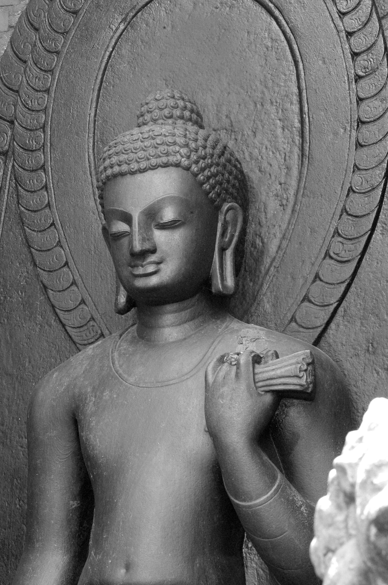 Black and white photograph of torso and head of Buddha holding implement in right hand at shoulder level