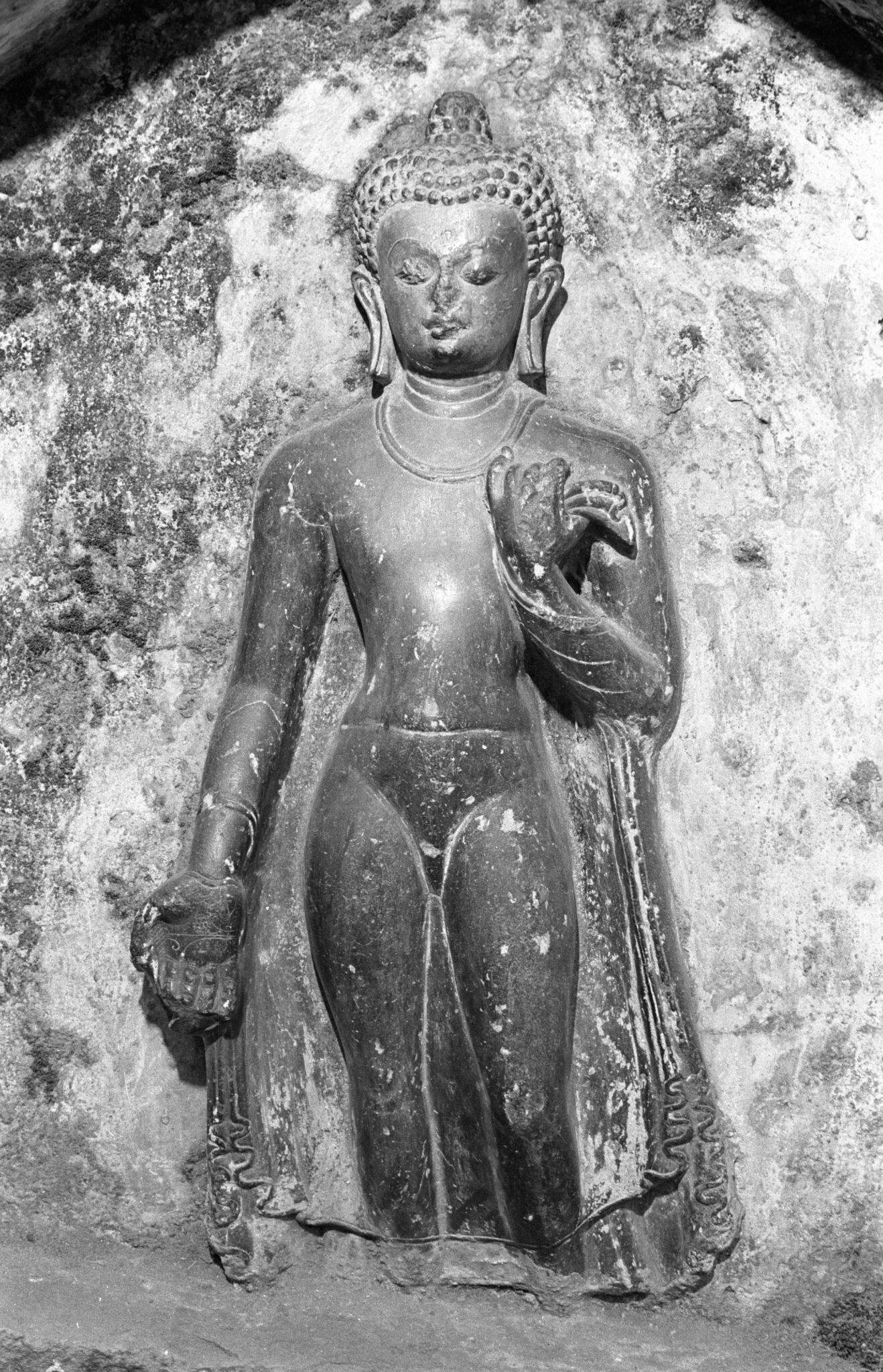Black and white photograph of relief sculpture depicting elegantly carved Buddha against rough-hewn background; feet are missing