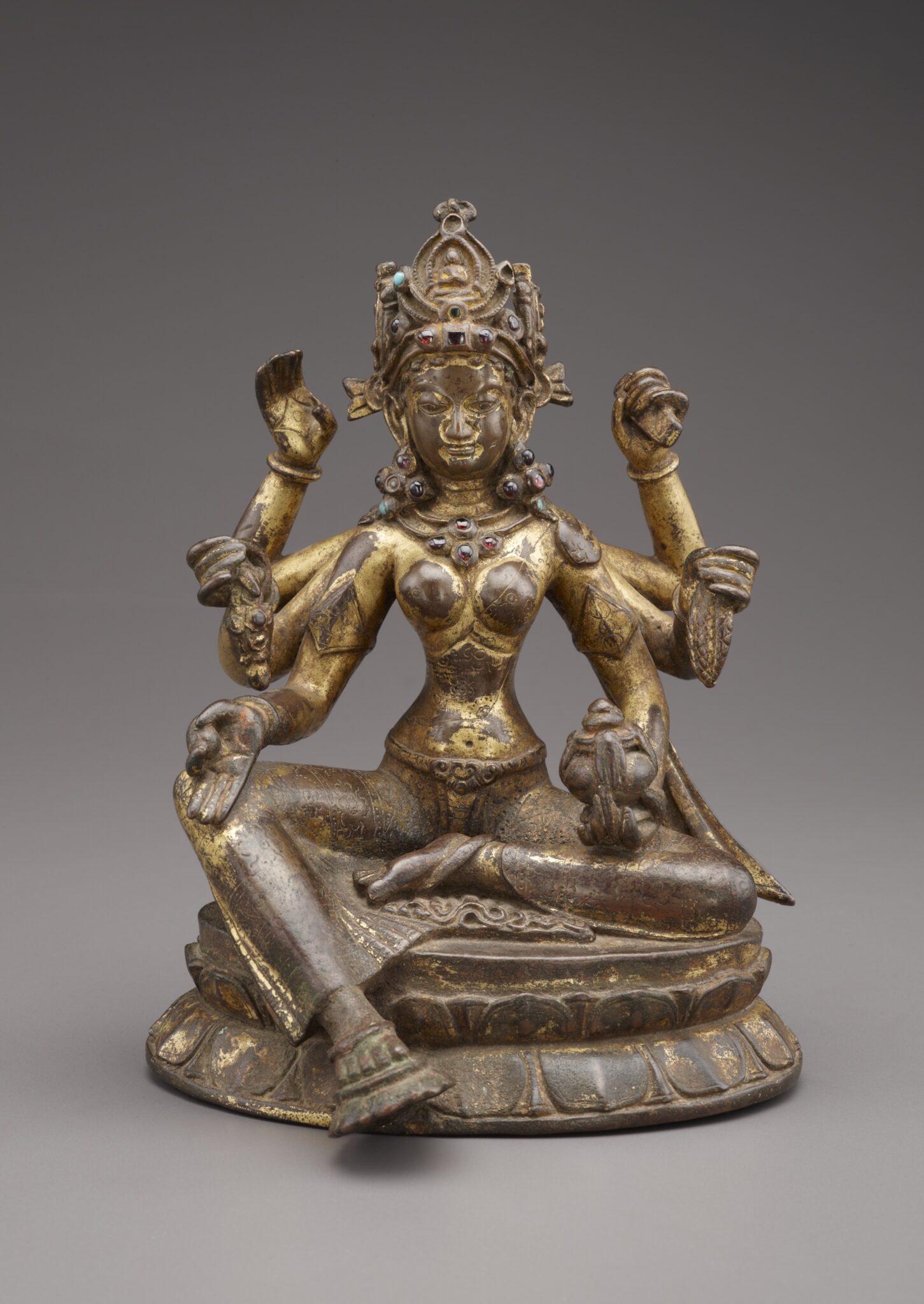 Tarnished gilded statuette depicting six-armed goddess seated on lotus pedestal; left foot rests on lotus blossom