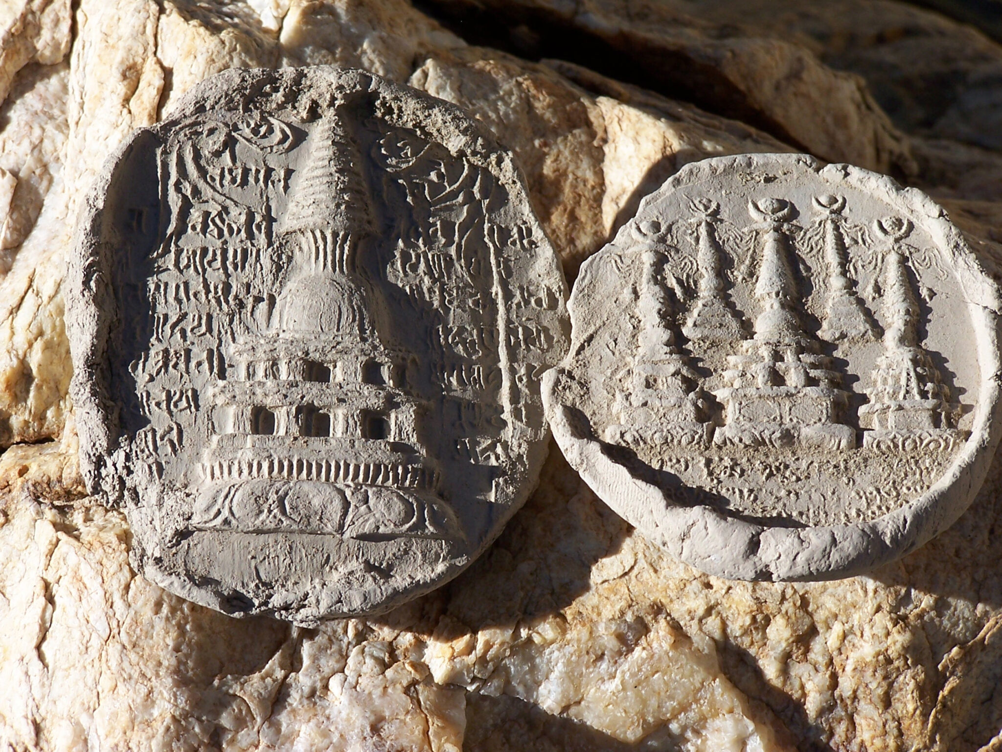 Two stamped clay discs arranged on beige rock: at left, one stupa amid text; at right, five stupas in two rows