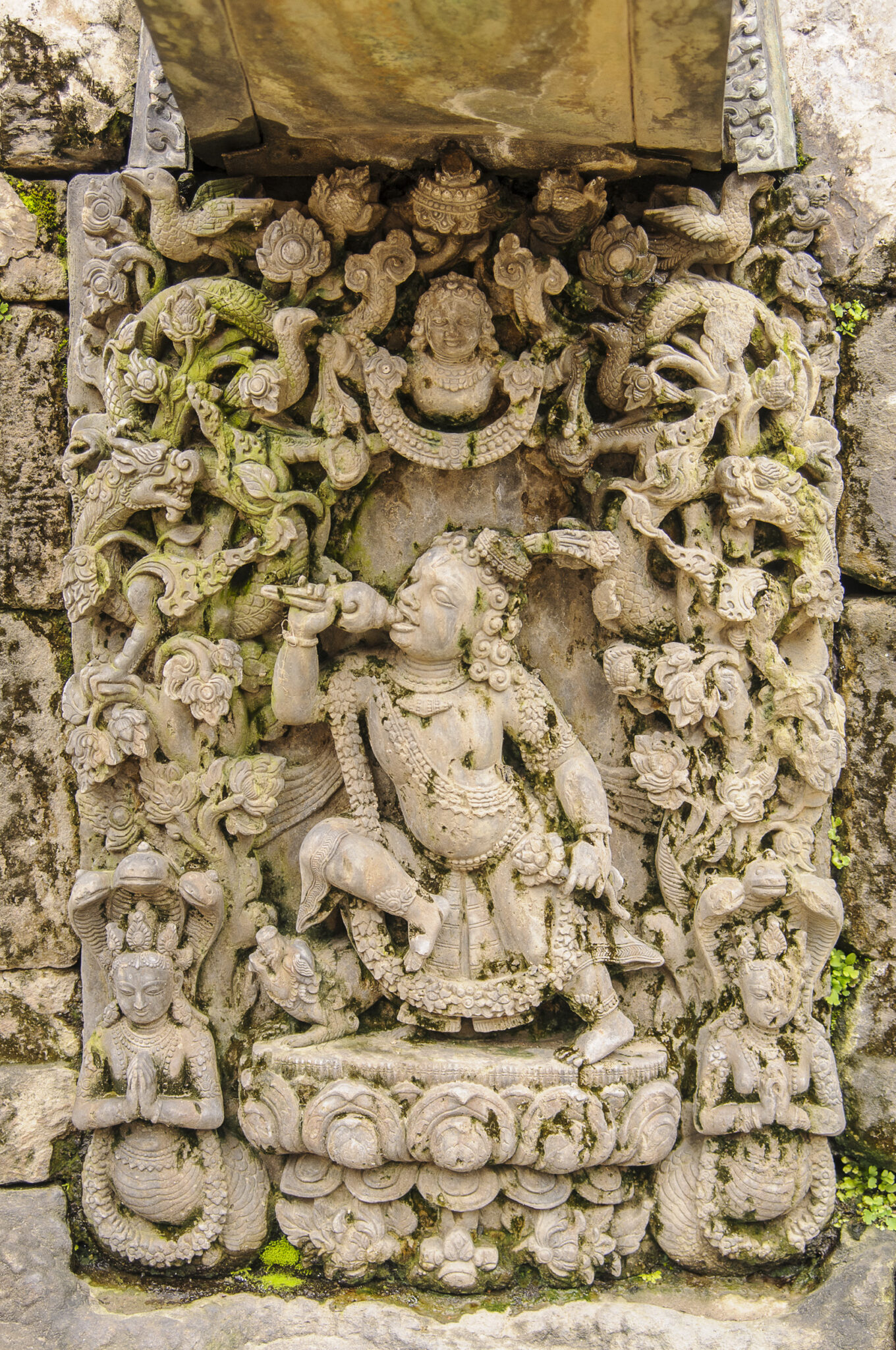 Relief sculpture depicting prince holding conch shell to mouth amidst twisting vines, blossoms, and deities