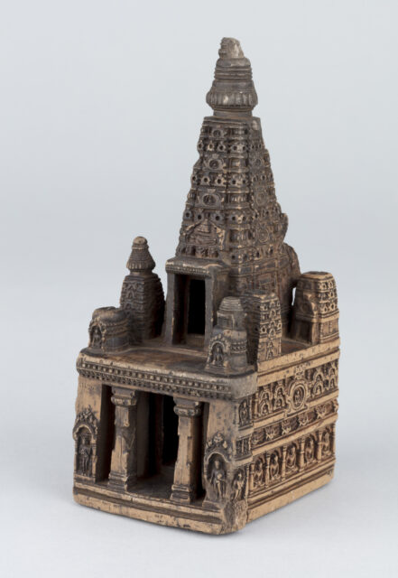 Miniature brown architectural model featuring square base with portico and slender tapered roof; decorated with intricately carved moldings