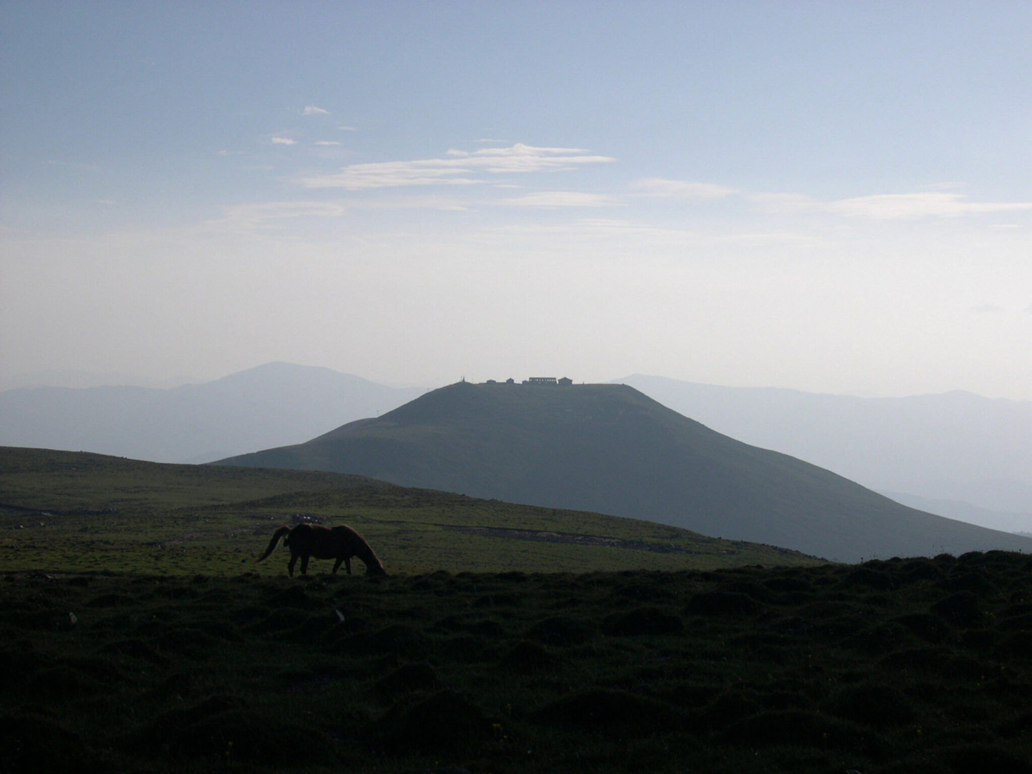 Wide view of mesa-topped mountain under hazy sky; lone horse grazes on green pasture in foreground