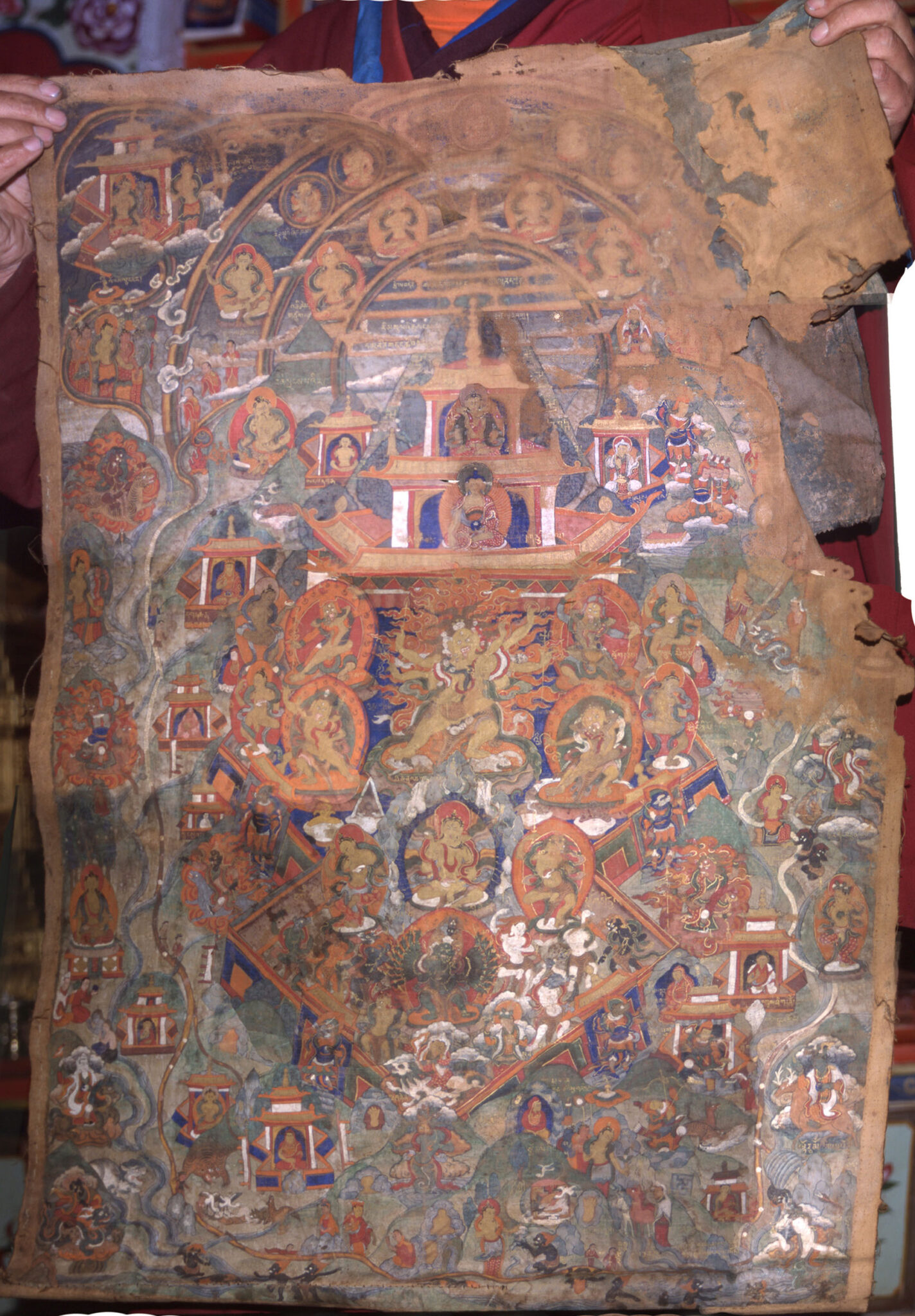 Tattered painting on textile depicting extravagant architectural structure covered in deity portraits amidst green landscape