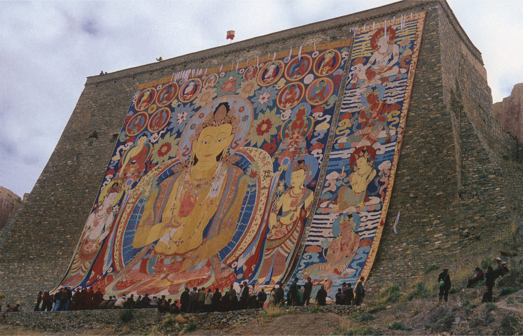 Brightly colored monumental textile depicting various deities hung on sloping stone wall; people standing at base indicate textile’s great size