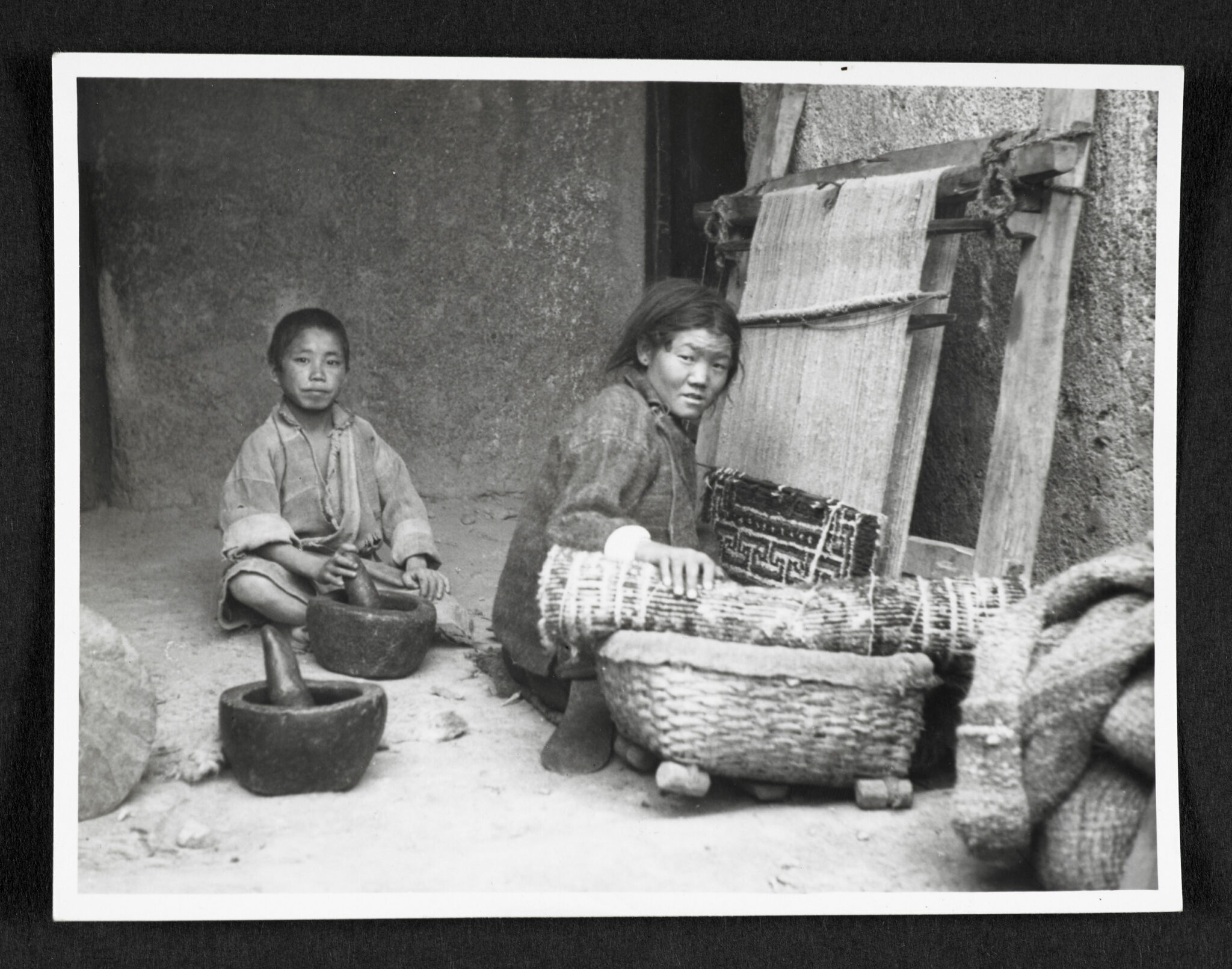 Black and white photograph of boy at left sitting behind mortar and pestle, and woman at right sitting at loom