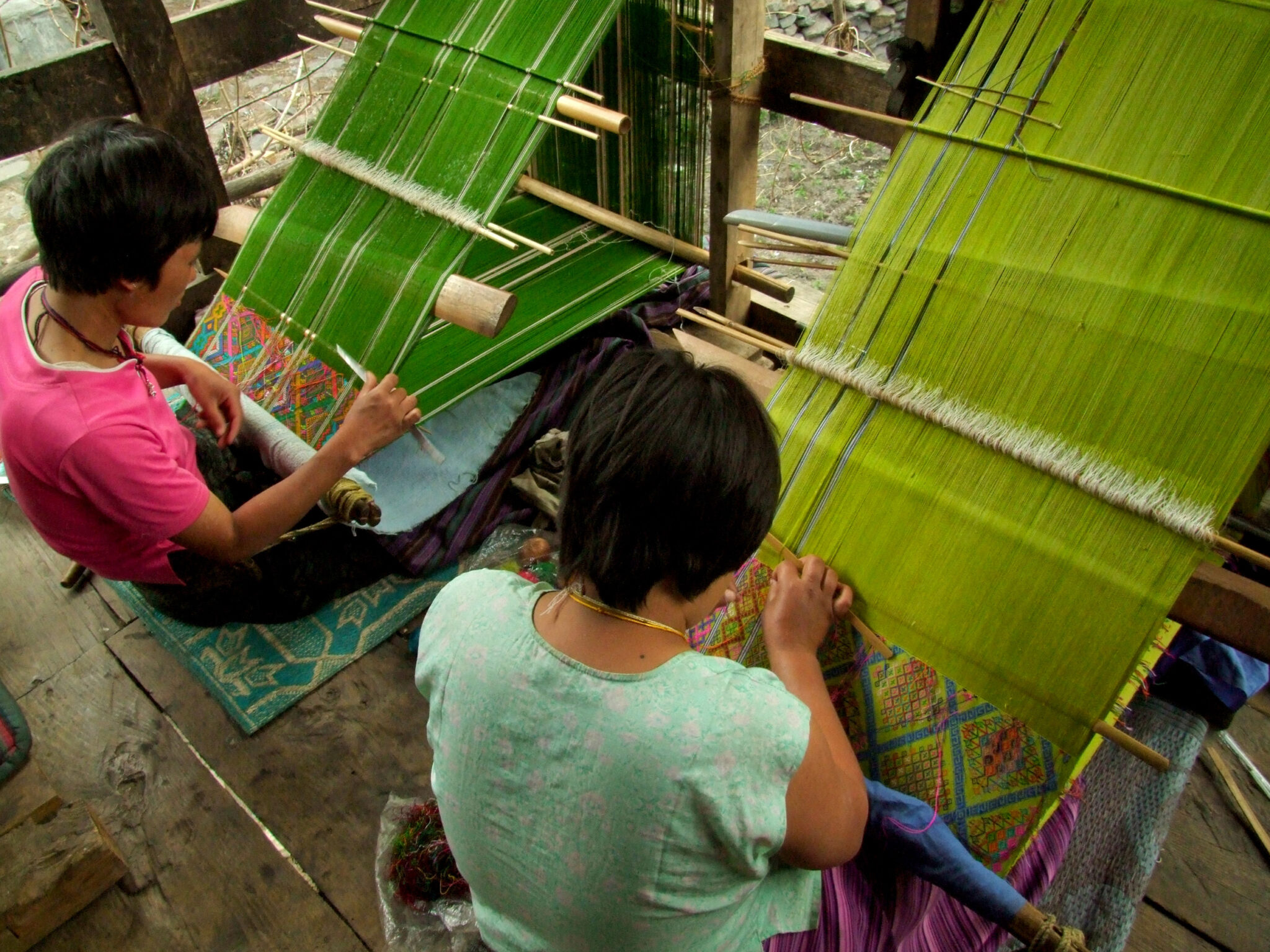 Two women seated on the floor before looms prepared with two different shades of green silk