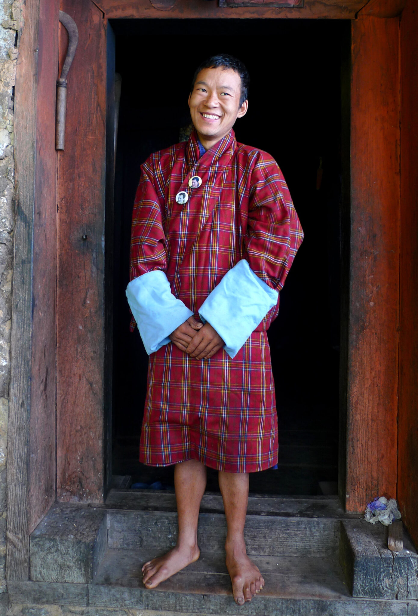 Man stands smiling in doorway wearing red plaid tunic with white cuffs covering forearms