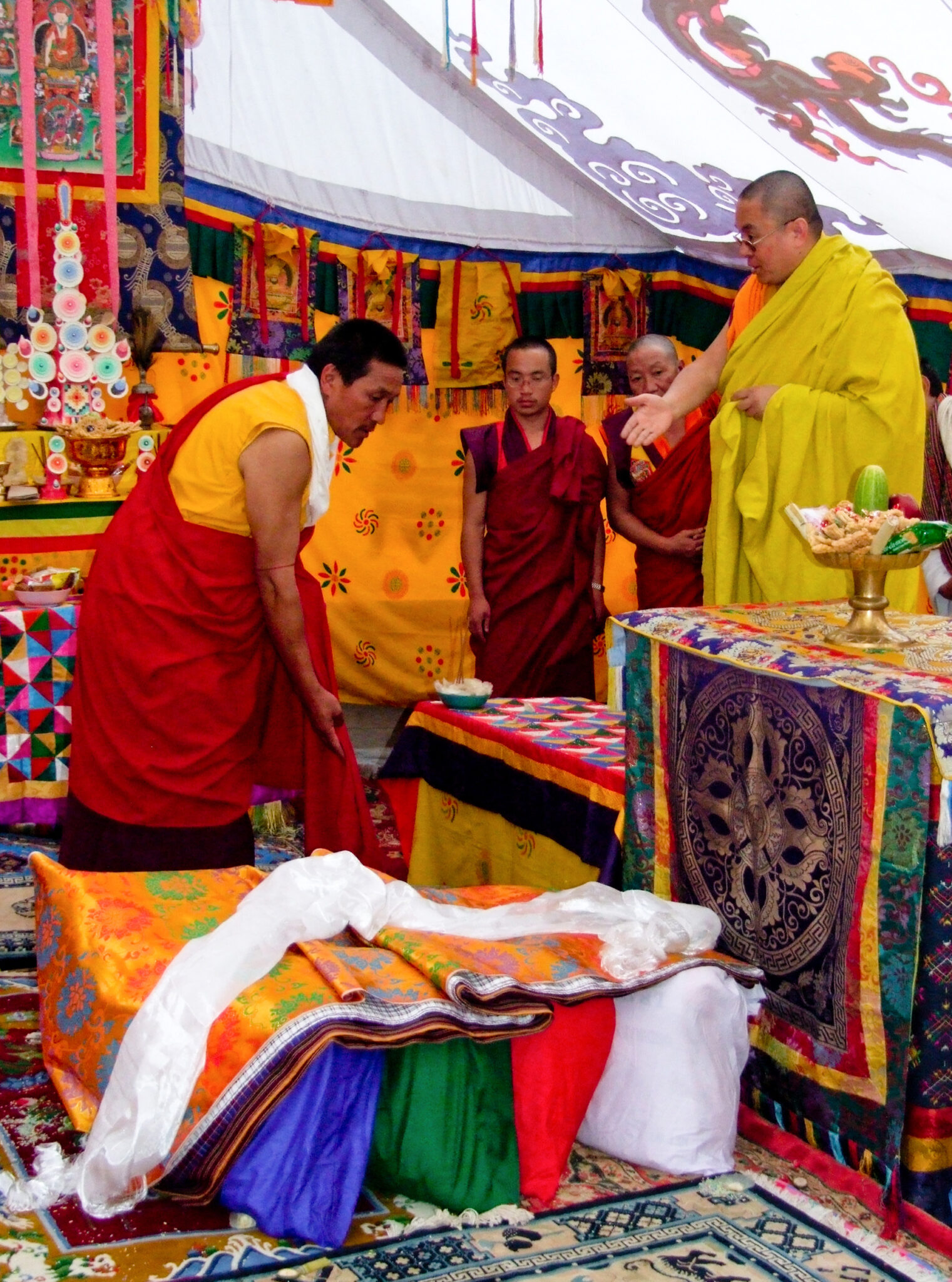 Abbott, in yellow robe, gestures to collection of textiles at base of altar in tent festooned with colorful textiles