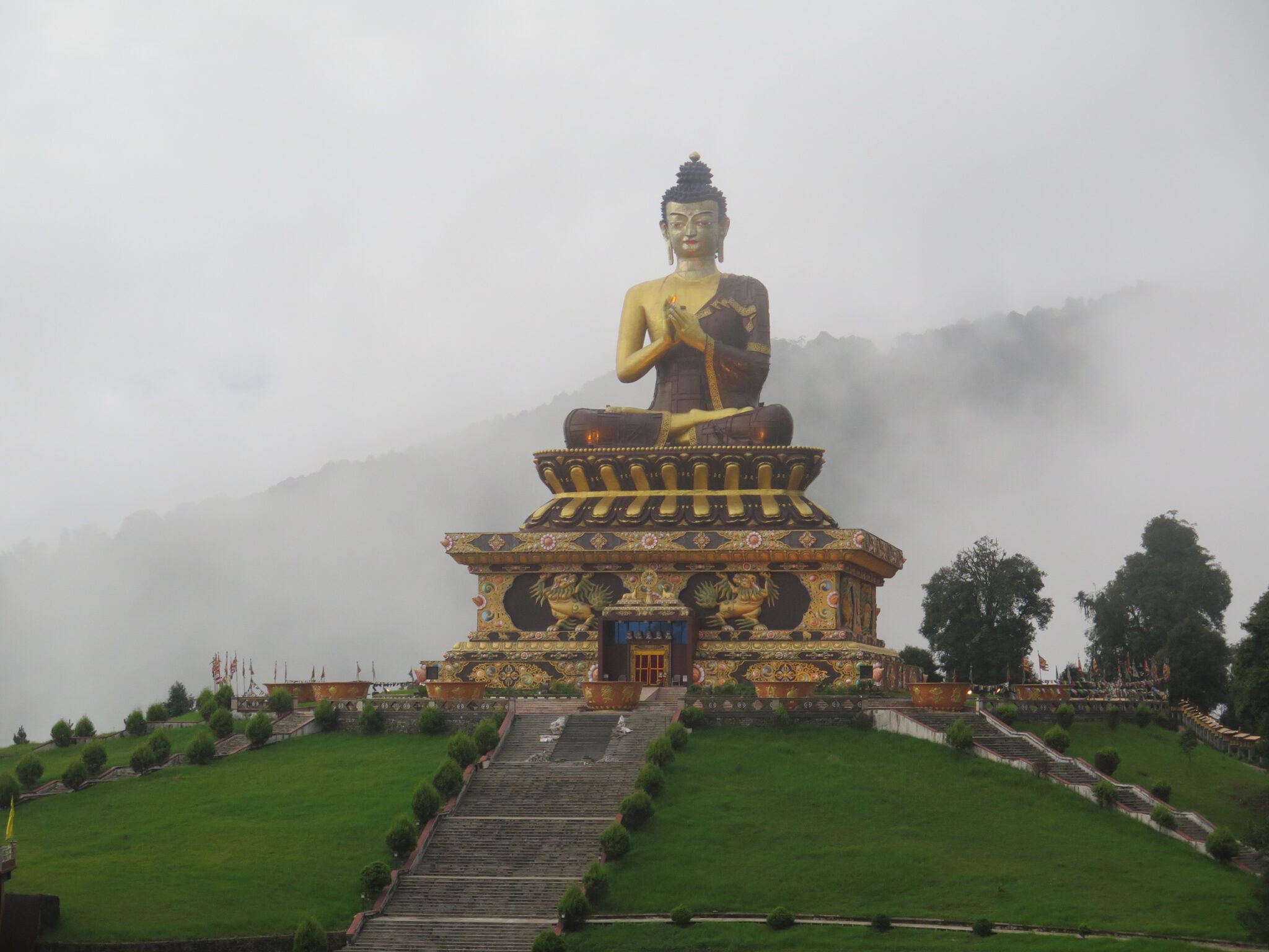 Colossal statue depicting golden Buddha seated atop plinth; situated on hilltop before clouded mountainside