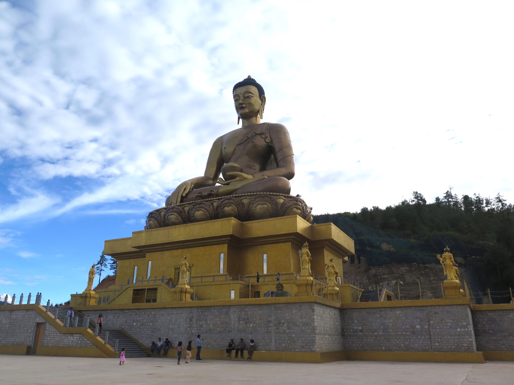 Colossal golden Buddha statue seated atop golden plinth, surrounded by terrace decorated with deity statues