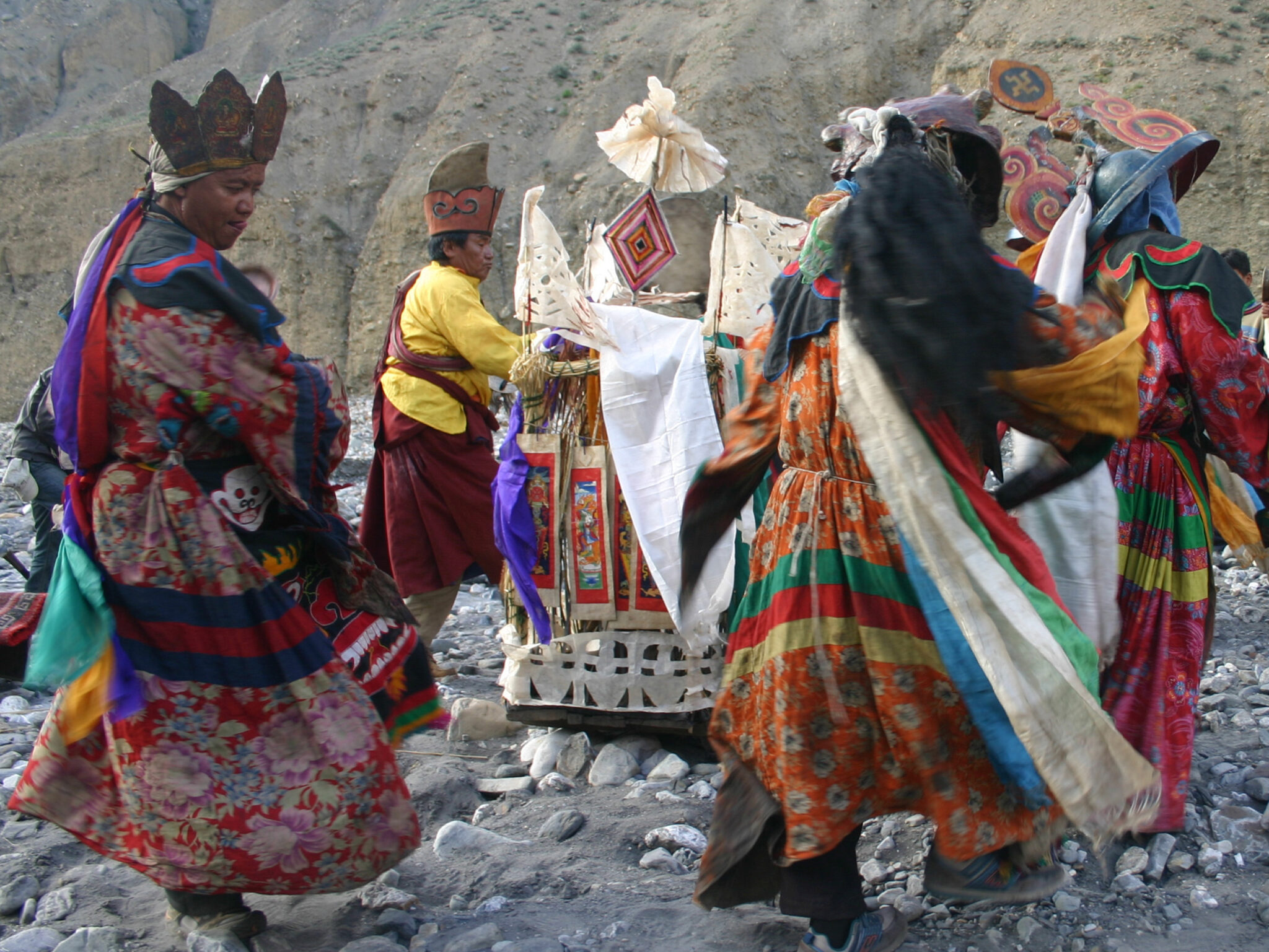 Lamas dressed in colorful robes dance around religious implement decorated with textiles and yarn lozenge