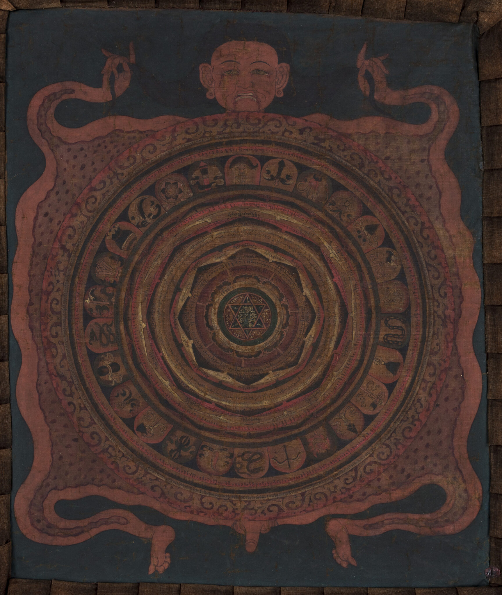 Brick-red mandala with lotus at center surrounded and supported by flayed body of figure holding his hair in his hands