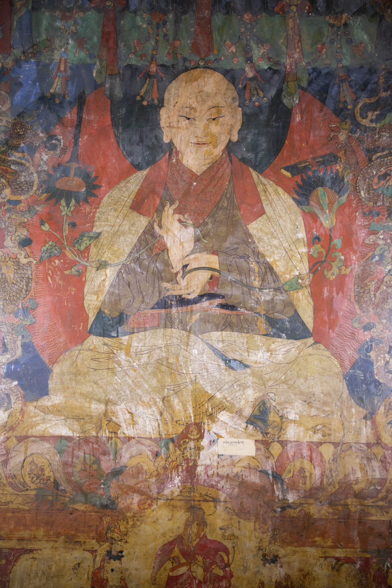 Seated spiritual leader with hands posed in mudras at chest holding two long-stemmed blossoms