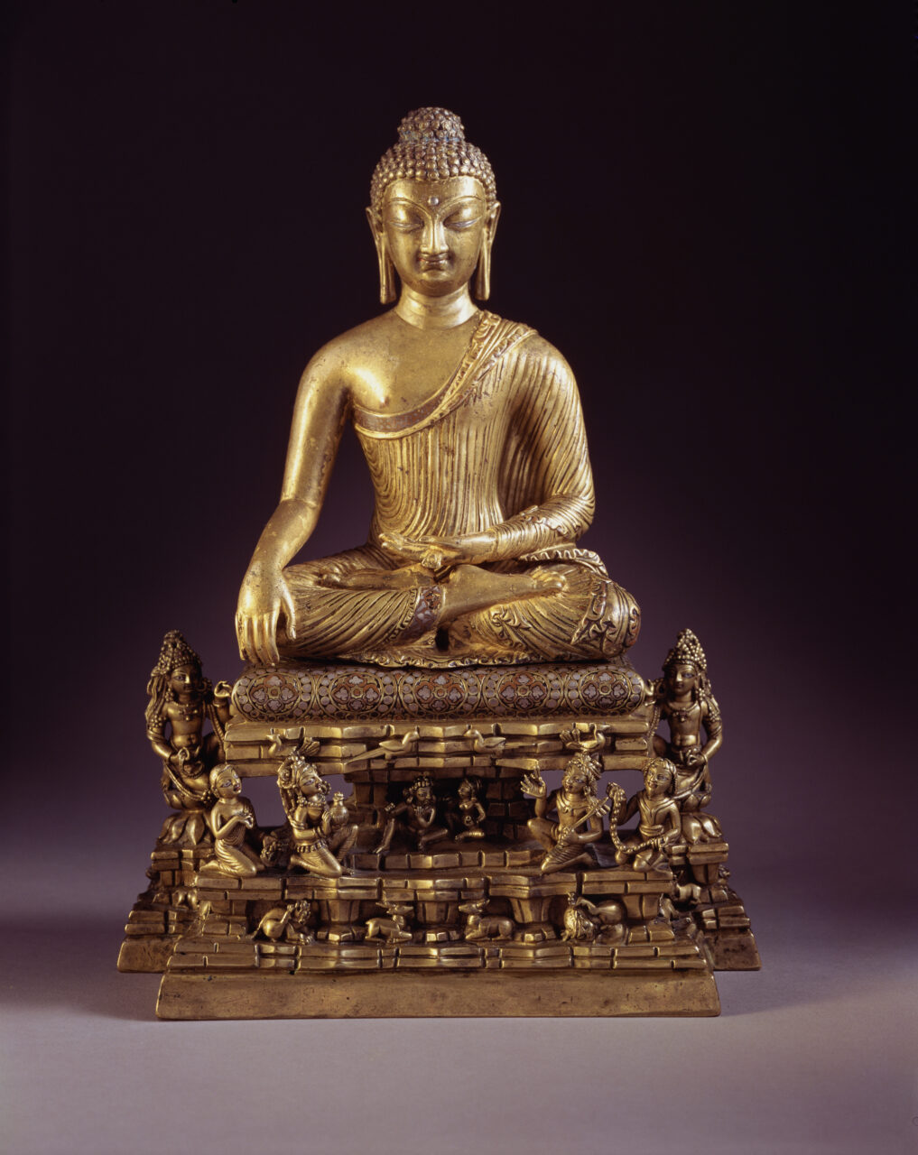 Bronze statuette of meditating Buddha seated on cushioned throne, around which many small figures kneel in reverence