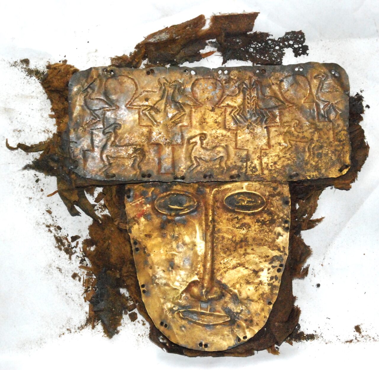 Golden burial mask with abstracted features wearing rectangular headdress depicting animal and architectural forms; it rests on decayed silk