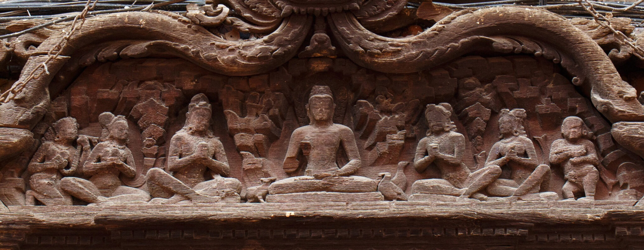 Medium view of densely carved brown wooden lintel beneath row of small windows; features scene with seven seated figures framed by scrollwork