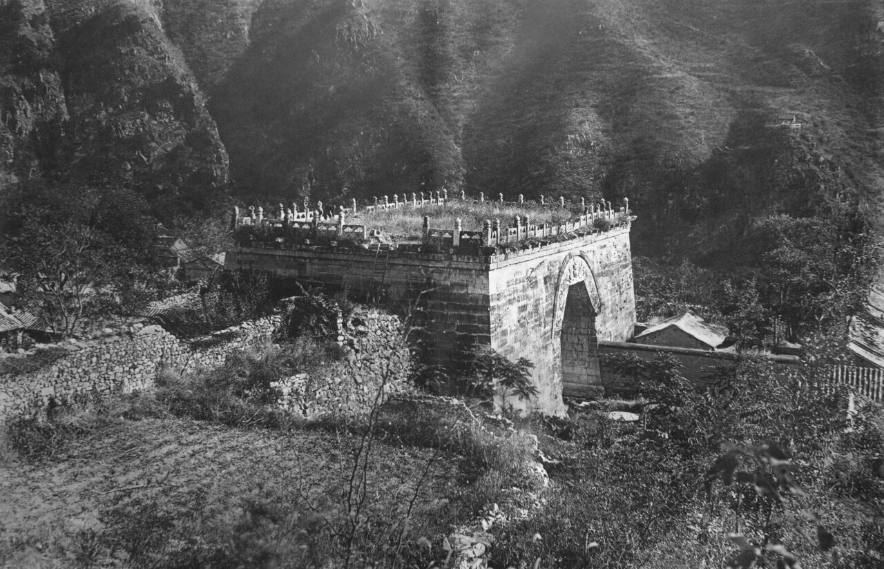Black and white photograph of arched stone structure overgrown with vegetation before scrub-covered mountainside