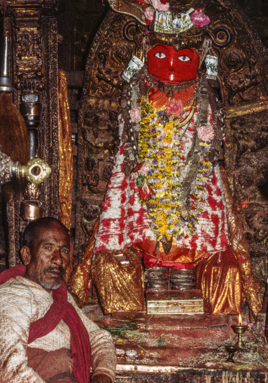 Man sits before statue adorned with garlands and textiles; statue has red, abstract face