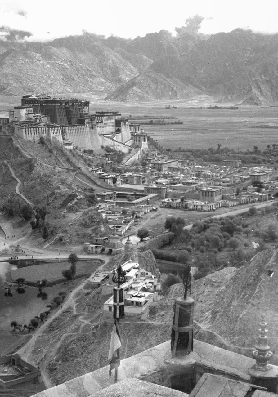 Black and white photograph of palace complex situated on peak of hill within mountain valley